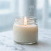 A lit candle in a plain glass jar on white marble, a thin thread of smoke rising