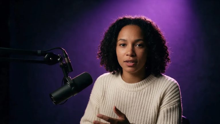 The same woman in the cream sweater speaking beside a podcast microphone under violet light